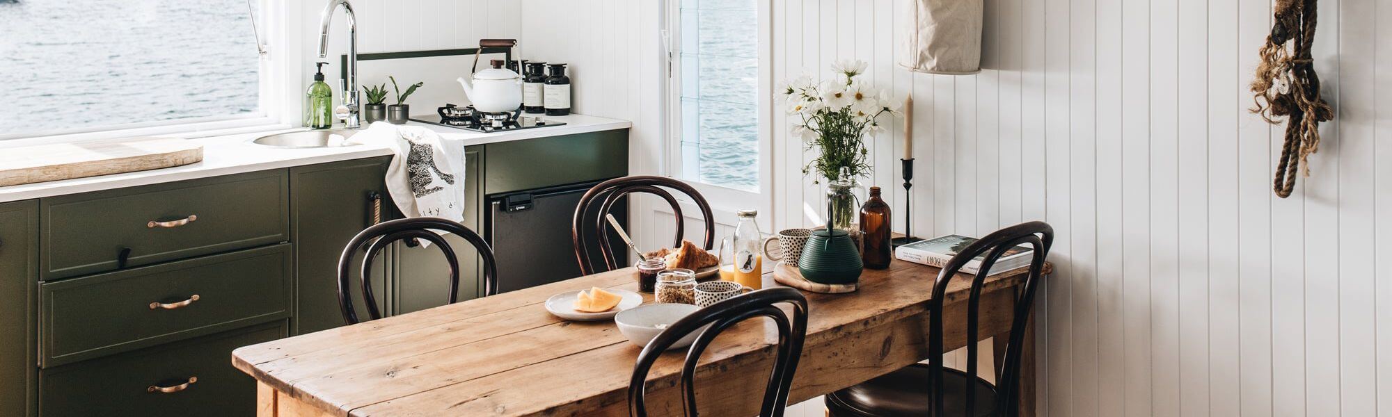Cozy waterfront kitchen with olive green cabinets, rustic wooden dining table, and natural light overlooking the water – Ren Studio.