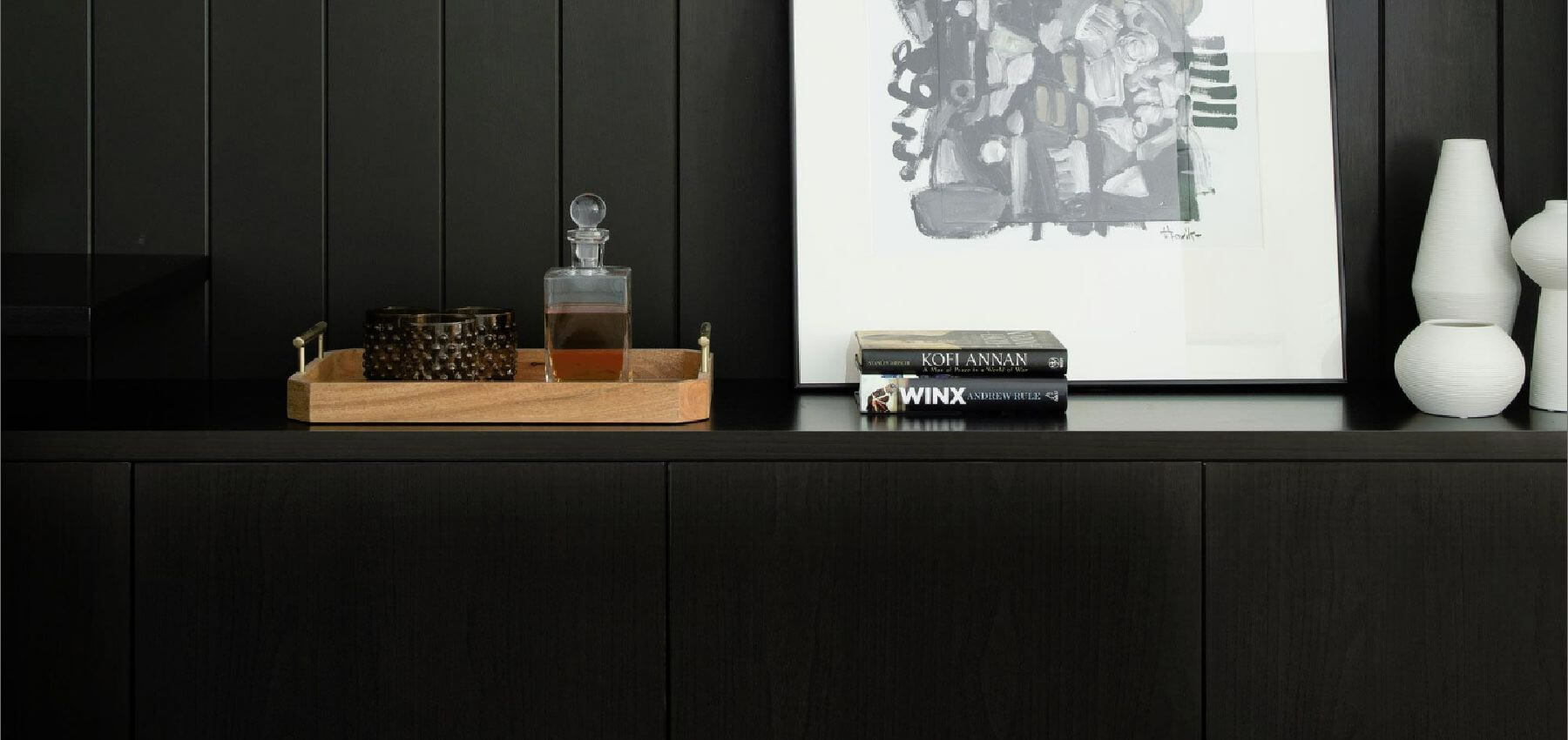 Black cabinetry with wooden tray, glass decanter, modern artwork, and white ceramic vases styled on a sleek sideboard – Ren Studio.