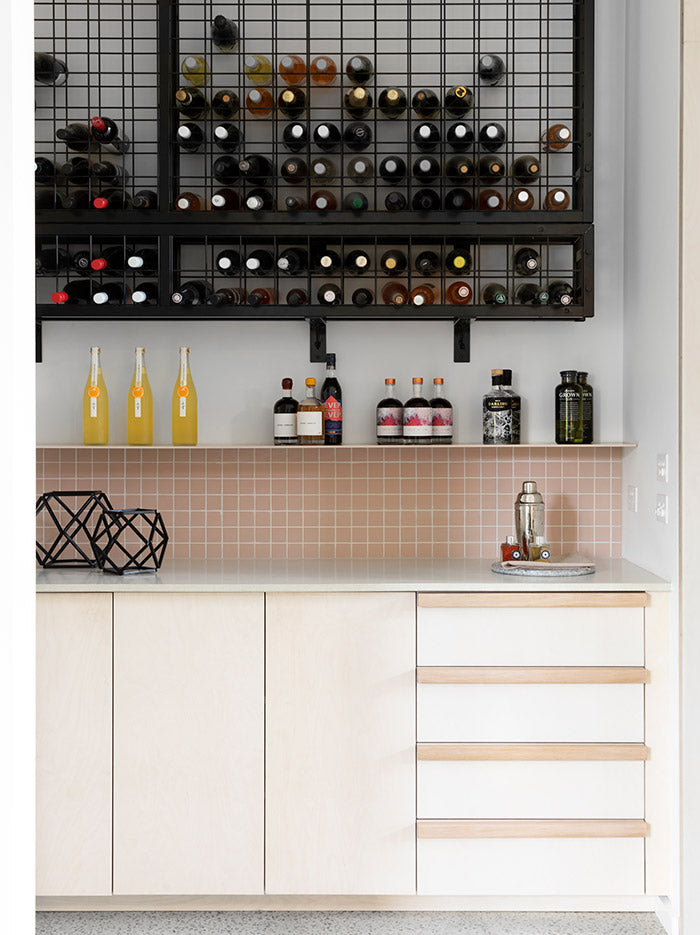 Modern home bar with light wood cabinetry, pink tile backsplash, wall-mounted wine rack, and assorted bottles on display – Ren Studio.